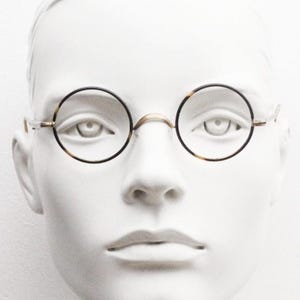 May include: Round eyeglasses with a thin gold bridge and black rims. The lenses are clear, and the frames have a tortoise shell pattern. The glasses are displayed on a white mannequin head, showcasing their design and style.