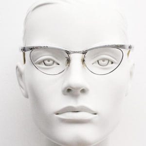 May include: Silver-framed cat-eye spectacles with clear lenses. The frames feature a decorative floral pattern along the top edge, displayed on a white mannequin head. The glasses have a classic, vintage aesthetic.