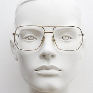 May include: Gold-rimmed square glasses with clear lenses on a white mannequin head.