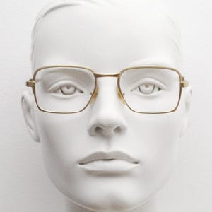 May include: A gold-toned metal frame with square lenses. The glasses are on a white mannequin head.