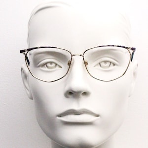 Könnte beinhalten: Eine Brille mit goldfarbenem Rahmen und einem gemusterten Rahmen in Lila und Blau. Die Brille hat eine Cat-Eye-Form und befindet sich auf einem weißen Mannequin-Kopf.