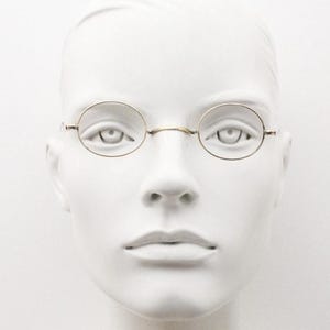 Peut inclure: Une paire de lunettes en métal doré dotée de montures circulaires fines et d'un pont simple. Les lunettes sont placées sur une tête de mannequin blanche, mettant en valeur le design minimaliste de la monture métallique et la construction délicate.
