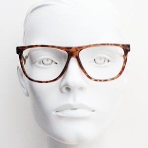 Puede incluir: Gafas de carey con un patrón moteado marrón y negro y una forma de montura rectangular. Las gafas cuentan con un puente fino y lentes transparentes, colocadas sobre una cabeza de maniquí blanca para mostrar la estructura del marco.