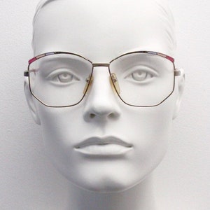 May include: A white mannequin head wearing gold-rimmed glasses with a multi-coloured trim. The glasses have a square shape with rounded corners.