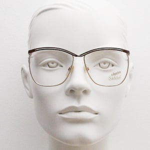 Könnte beinhalten: Eine Brille mit goldfarbenem Rahmen und braunen Bügeln. Die Brille hat eine Cat-Eye-Form und der Markenname "elastica Safilo" ist auf der rechten Linse aufgedruckt.