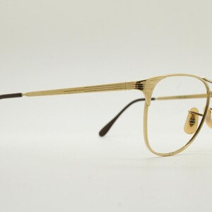 Puede incluir: Gafas de metal doradas con patillas marrones. Las gafas tienen una forma rectangular redondeada.