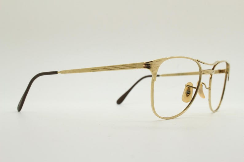 Puede incluir: Gafas de metal doradas con patillas marrones. Las gafas tienen una forma rectangular redondeada.