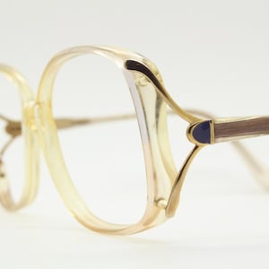 May include: Vintage eyeglasses with a light yellow frame and gold-coloured accents. The temples have a brown and gold pattern with a small blue detail. The glasses have a unique, oversized shape.