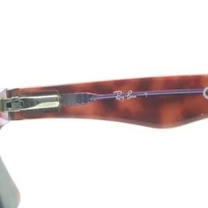 May include: Close-up of the hinge and temple arm of a pair of tortoiseshell eyeglasses. The temple arm is inscribed with "Ray-Ban" and "CE".