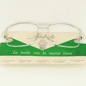 May include: Silver-framed eyeglasses with a double bridge design rest on a vintage tag. The tag features green and cream colors with Italian text, including "La moda con la nuova linea."