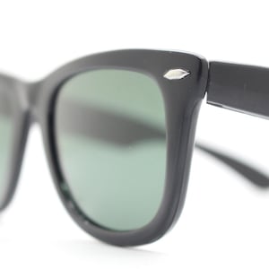 Puede incluir: Gafas de sol negras con un dise&ntilde;o cl&aacute;sico. Las monturas son gruesas y angulares, con un sutil acento plateado en las patillas. Las lentes son de un verde apagado, ofreciendo un contraste con las monturas oscuras.