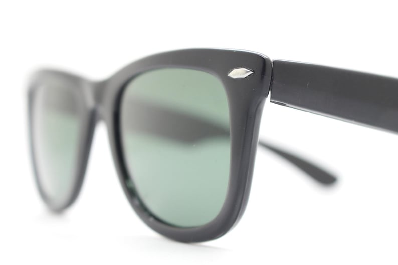 Puede incluir: Gafas de sol negras con un dise&ntilde;o cl&aacute;sico. Las monturas son gruesas y angulares, con un sutil acento plateado en las patillas. Las lentes son de un verde apagado, ofreciendo un contraste con las monturas oscuras.