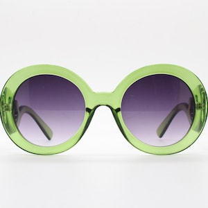 May include: Round sunglasses featuring a translucent lime green plastic frame and dark purple gradient lenses. The circular frames are thick and bold, with matching green temples visible behind the lenses, creating a cohesive look.