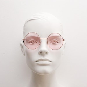 May include: Pink-tinted, octagonal sunglasses with a gold-colored metal frame. The glasses are displayed on a white mannequin head, showcasing the eyewear's shape and color. The lenses are a soft pink, offering a stylish and unique look.