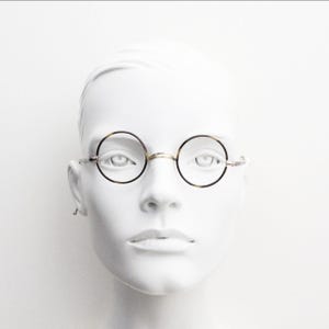 May include: Round spectacles featuring a thin, tortoiseshell-patterned frame with a metallic bridge and slender temples. The circular lenses are clear, and the frame design highlights a classic, minimalist aesthetic with a focus on the circular shape.