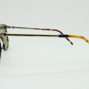 May include: Tortoiseshell sunglasses with gold metal arms and a black accent on the temple. The sunglasses have green lenses and say "MARC JACOBS" on the arm.