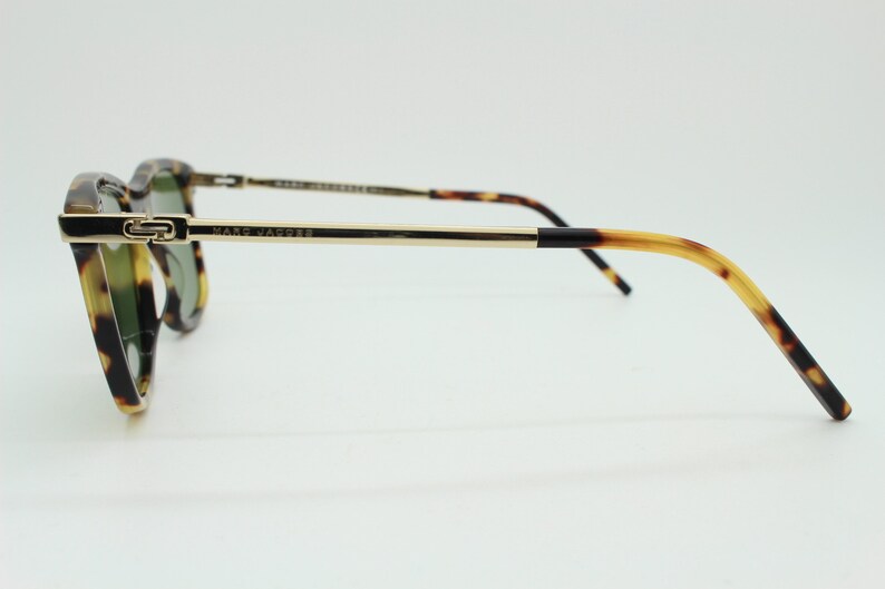 May include: Tortoiseshell sunglasses with gold metal arms and a black accent on the temple. The sunglasses have green lenses and say "MARC JACOBS" on the arm.