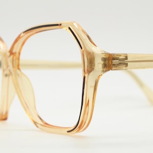May include: Close-up of vintage eyeglasses with a light amber-colored frame. The glasses have a large, angular shape with a subtle, translucent quality. The temples and bridge are a similar hue, creating a cohesive design.