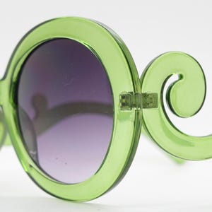 May include: Lime green sunglasses featuring large, circular frames and distinctive, curved, scroll-like temples. The lenses are dark and gradient, transitioning from a deeper shade at the top to a lighter tint toward the bottom of the circular rims.