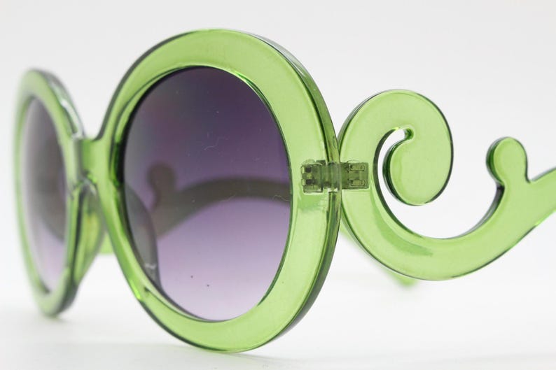 May include: Lime green sunglasses featuring large, circular frames and distinctive, curved, scroll-like temples. The lenses are dark and gradient, transitioning from a deeper shade at the top to a lighter tint toward the bottom of the circular rims.
