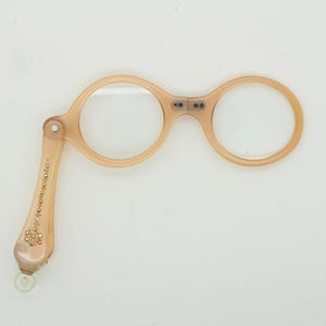Op de afbeelding: Een perzikroze leesbril met ronde glazen en een opvouwbaar handvat. Het handvat is versierd met kleine, decoratieve strass-steentjes. De bril staat tegen een effen witte achtergrond.
