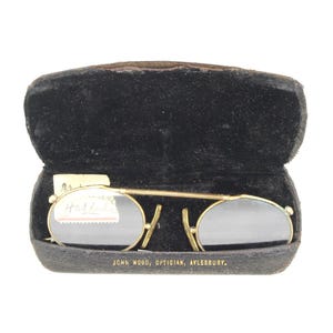 May include: Vintage gold-tone clip-on sunglasses with oval lenses, resting in a black velvet case. The case is open, revealing the glasses and a small tag. The case is labeled "JOHN WOOD, OPTICIAN, AYLESBURY."