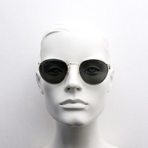 Pu&ograve; includere: Occhiali da sole rotondi con montatura dorata e lenti scure. Le montature sono sottili e metalliche. Gli occhiali sono esposti su una testa di manichino bianca su sfondo bianco. Le lenti sono di colore scuro.