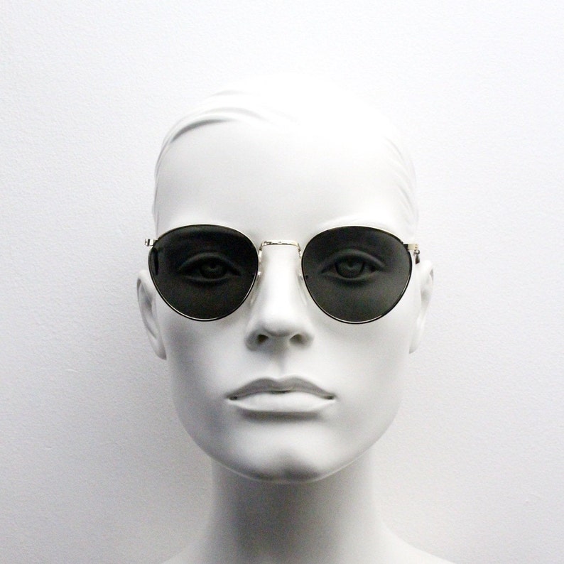 Pu&ograve; includere: Occhiali da sole rotondi con montatura dorata e lenti scure. Le montature sono sottili e metalliche. Gli occhiali sono esposti su una testa di manichino bianca su sfondo bianco. Le lenti sono di colore scuro.