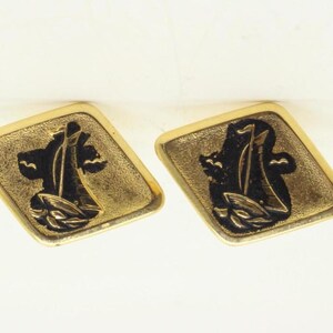 Könnte beinhalten: Ein Paar Manschettenknöpfe aus goldfarbenem Metall, jeweils mit einer rautenförmigen Vorderseite und einer vertieften schwarzen Mitte. Die Mitte zeigt ein erhabenes, detailliertes Relief eines Segelboots auf Wellen mit strukturierter Oberfläche.