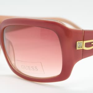 May include: Rose-colored sunglasses with a glossy finish and gradient lenses. The frame features a gold-tone metal accent on the temple with a stylized G logo flanked by two small clear crystals. The word GUESS is printed on the right lens.