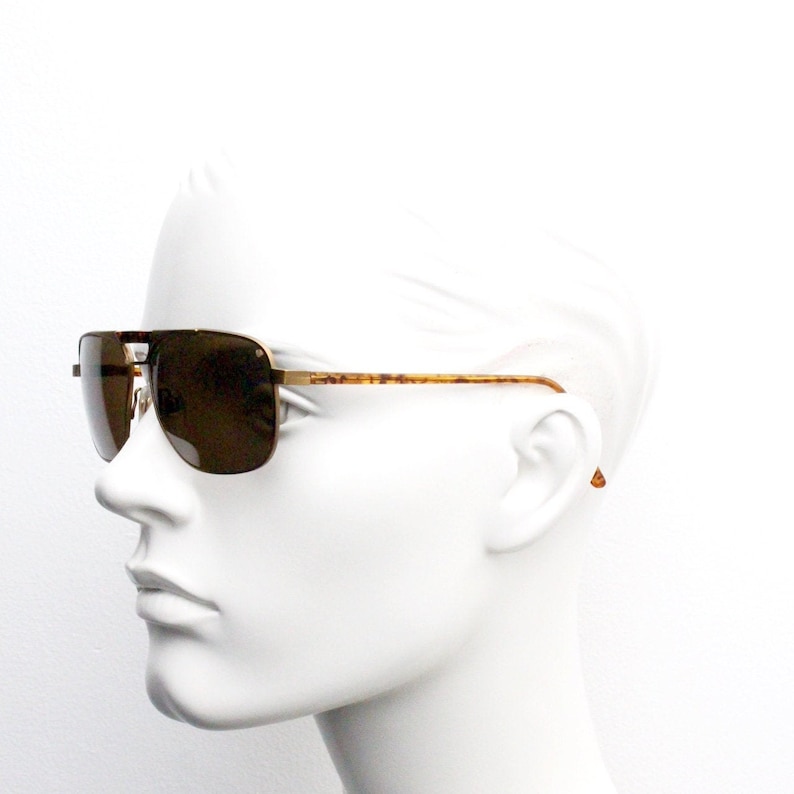 May include: A pair of gold-tone aviator sunglasses with brown lenses. The sunglasses are on a white mannequin head.