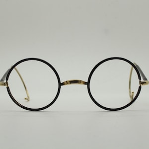 May include: Round eyeglasses with black frames and gold-colored accents. The lenses are clear, and the bridge and temples are a contrasting gold color. The glasses have a classic, vintage aesthetic.