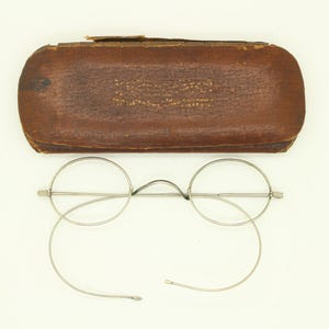 Peut inclure: Lunettes rondes anciennes avec une monture en métal argenté et des verres transparents, sous un étui à lunettes en cuir marron. L'étui porte un texte en relief. Les lunettes ont une esthétique classique et vintage.