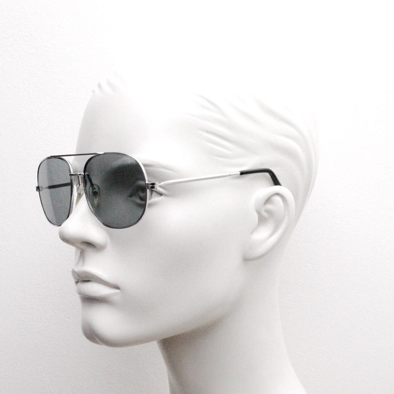 May include: Silver aviator sunglasses with dark gray lenses. The sunglasses are on a white mannequin head.