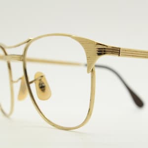 Puede incluir: Gafas de metal doradas con montura redonda y puente fino y recto. Las patillas son finas y tienen un dise&ntilde;o sutilmente texturizado.