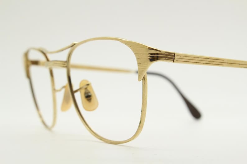 Puede incluir: Gafas de metal doradas con montura redonda y puente fino y recto. Las patillas son finas y tienen un dise&ntilde;o sutilmente texturizado.