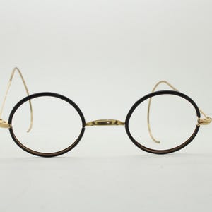 May include: Round spectacles featuring thin, dark brown circular rims and a metallic gold bridge. The temples are constructed from slender, curved gold wire designed to hook behind the ears, providing a lightweight and minimalist frame structure.