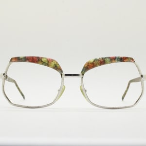 Könnte beinhalten: Brille mit silbernem Rahmen und farbenfrohem Blumenmuster an den oberen Rändern. Die Gläser sind klar, und die Fassungen haben eine geometrische, übergroße Form. Die Bügel sind dünn und silberfarben, mit einer leichten Krümmung.