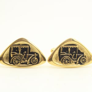 Könnte beinhalten: Ein Paar goldfarbene Manschettenknöpfe in dreieckiger Form. Jeder Manschettenknopf verfügt über ein vertieftes schwarzes Mittelfeld mit einem detaillierten Reliefdesign eines Oldtimers, das eine klassische Autosilhouette mit Rädern zeigt.