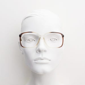 May include: A pair of spectacles featuring a rectangular frame with a gradient design that transitions from a dark brown tortoiseshell pattern at the top to a clear, transparent finish at the bottom, resting on a white mannequin head.