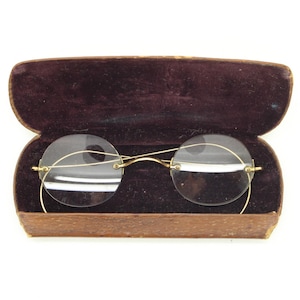 Peut inclure: Une paire de lunettes rondes sans monture avec des cadres fins en métal doré et un pont incurvé, reposant dans un étui rectangulaire ouvert à la texture brune et doublé de velours violet foncé. Les lunettes ont des verres clairs et des branches fines.