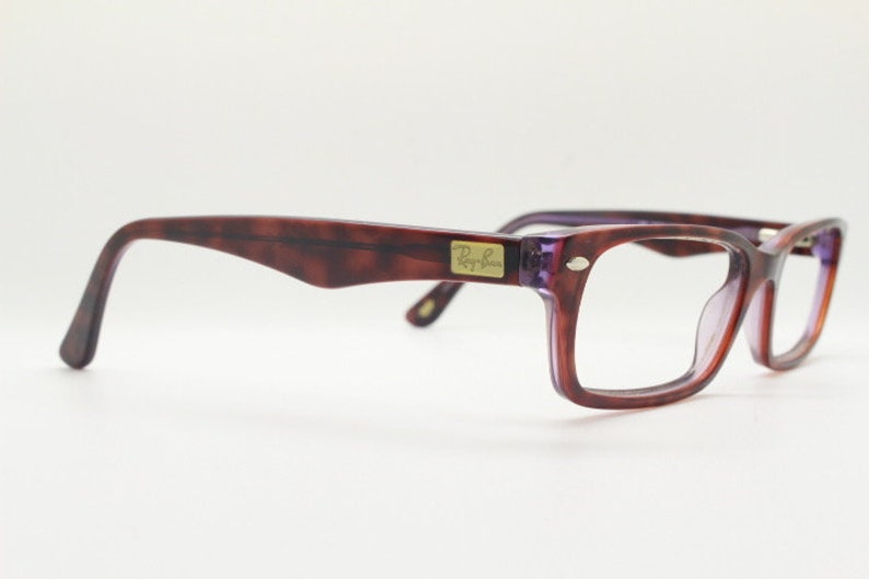 May include: A pair of tortoiseshell eyeglasses with purple frames and clear lenses. The Ray-Ban logo is visible on the right temple.