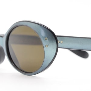 Może przedstawiać: Okulary przeciwsłoneczne w kształcie owalu, w kolorze niebieskim, z brązowymi szkłami. Okulary mają metalową oprawkę i są zaprojektowane w stylu retro.