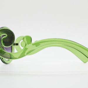May include: Translucent lime green sunglasses featuring a distinctive swirling, scroll-like design on the temples. The frames are made of a smooth, glossy material with dark tinted lenses visible from the side profile view.