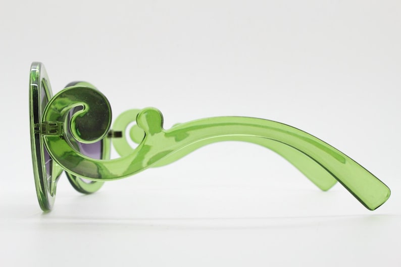 May include: Translucent lime green sunglasses featuring a distinctive swirling, scroll-like design on the temples. The frames are made of a smooth, glossy material with dark tinted lenses visible from the side profile view.