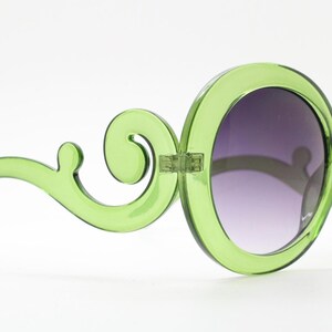 May include: Lime green sunglasses featuring large, circular frames and distinctive, swirling, scroll-like arms. The lenses are dark and have a gradient tint, transitioning from a darker shade at the top to a lighter shade at the bottom.
