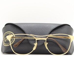 Puede incluir: Gafas con montura dorada y estuche de cuero negro. El estuche tiene el logotipo de Ray-Ban con el texto "Totally UV protected. Guaranteed by Luxottica."