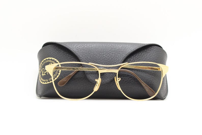Puede incluir: Gafas con montura dorada y estuche de cuero negro. El estuche tiene el logotipo de Ray-Ban con el texto "Totally UV protected. Guaranteed by Luxottica."