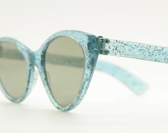 Könnte beinhalten: Eine Sonnenbrille im Cat-Eye-Stil in hellblau mit glitzerndem Finish. Die Sonnenbrille hat einen dünnen, hellblauen Rahmen und graue Gläser.
