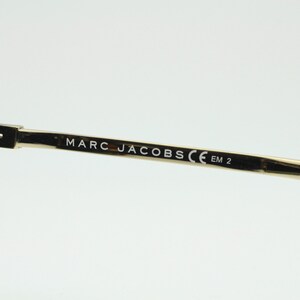 May include: Close-up of the arm of a pair of tortoiseshell sunglasses with gold metal arms. The arm is engraved with the text "MARC JACOBS CE EM 2".